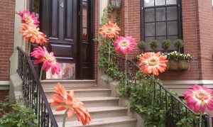 Eight oversized, fake orange and pink flowers line the railing leading to a lovely black Back Bay door 